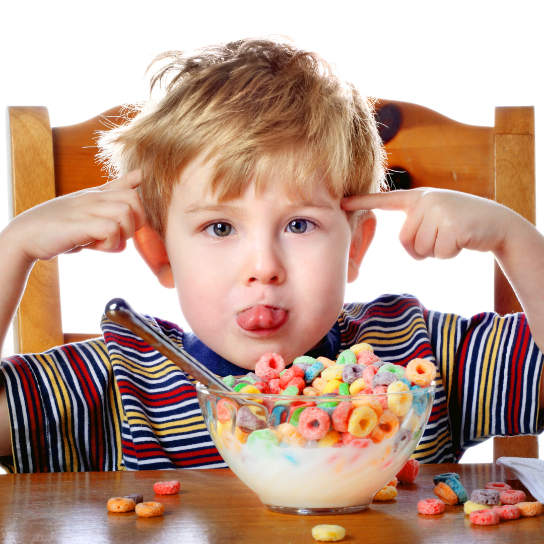 Junge sitzt vor einer Schüssel bunter Frühstückscerealien, streckt frech die Zunge heraus und hält sich mit beiden Zeigefingern die Ohren zu – symbolisch für Überdrehtheit nach zucker- und farbstoffreichen Lebensmitteln.